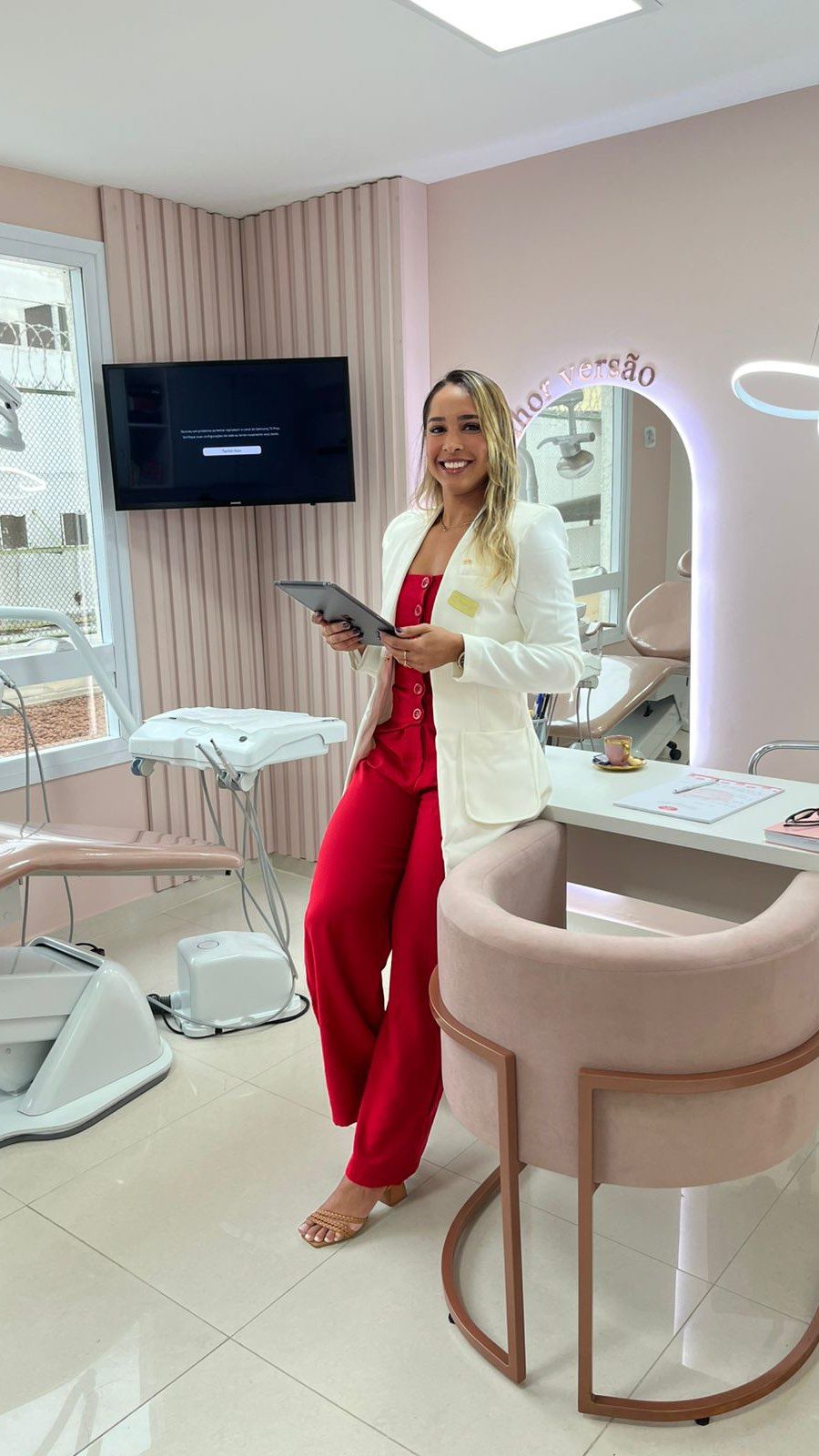 Dra. Dyene Oliveira em pé no consultório em Bangu, com paredes em rosa claro e cadeira odontológica.